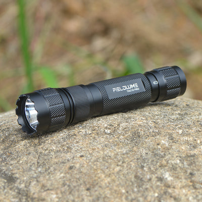 Black flashlight with 'FIELDLUME' branding on a stone surface with a blurred natural background