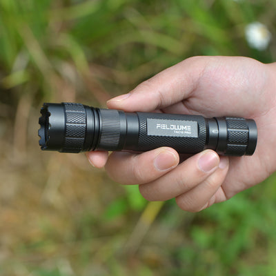 Hand holding a black flashlight with 'FIELDLUME' branding against a blurred natural background